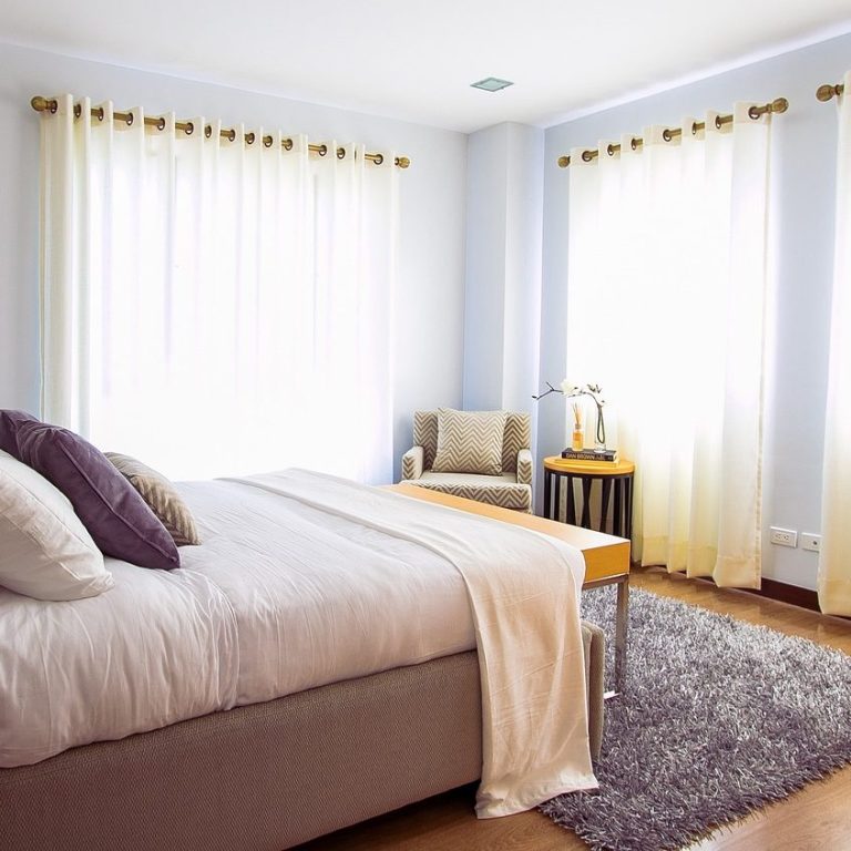 Raum und Heimtextilien Helles Schlafzimmer mit Bett, Sessel und großen Fenstern, die Tageslicht hereinlassen.